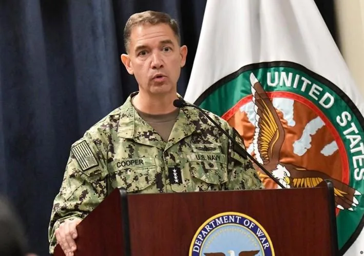 Oficial de la Marina de EE. UU. con uniforme de camuflaje y placa "COOPER" hablando en un podio. Al fondo, una bandera con el emblema de "United States".