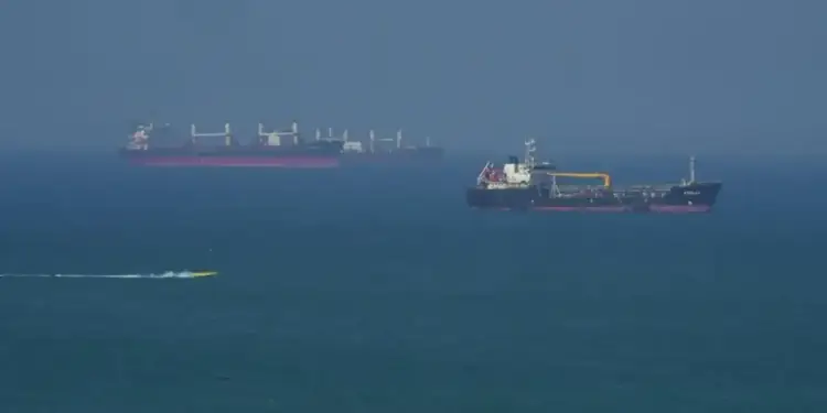 Vista aérea de buques de carga y un tanquero en el mar, con una pequeña embarcación amarilla en primer plano. Representa el comercio marítimo global.