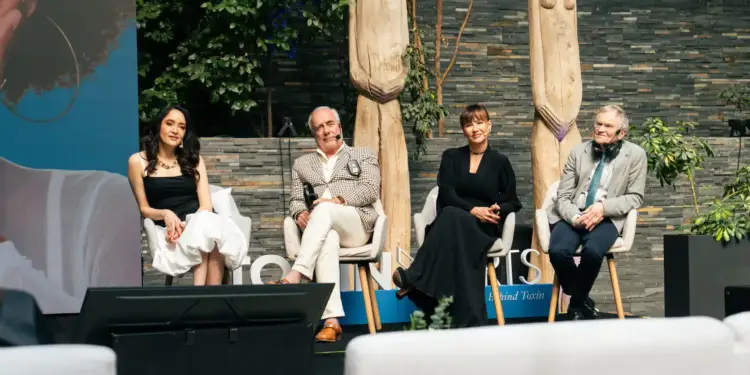 Cuatro panelistas sentados en sillones blancos durante un evento profesional al aire libre con esculturas de madera de fondo.
