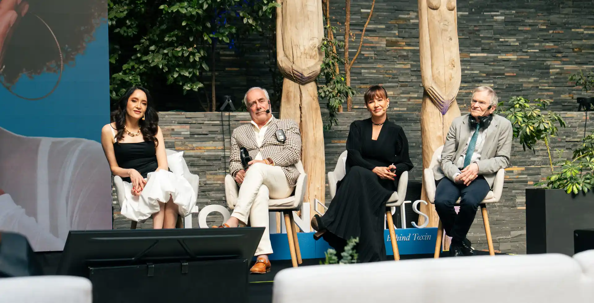 Cuatro panelistas sentados en sillones blancos durante un evento profesional al aire libre con esculturas de madera de fondo.
