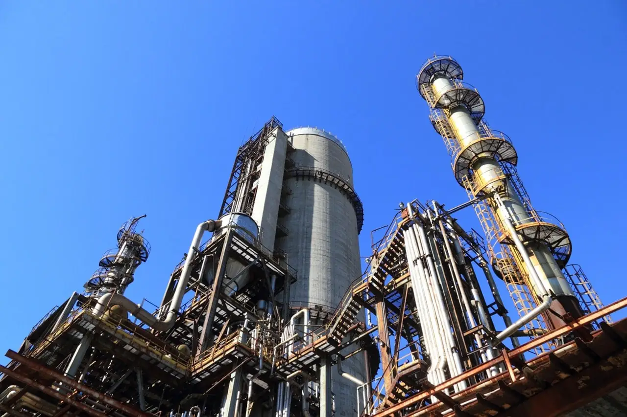Vista contrapicado de una planta industrial con torres de procesamiento, tuberías y estructuras metálicas bajo un cielo azul.