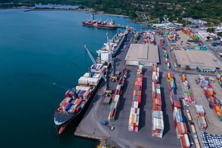 Vista aérea de un puerto con buques de carga atracados, grúas y extensas filas de contenedores de colores en la terminal.