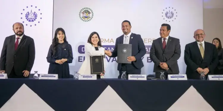 Grupo de seis ejecutivos en una ceremonia de firma de acuerdo. Dos personas centrales intercambian documentos y se estrechan la mano. Fondo con emblemas.
