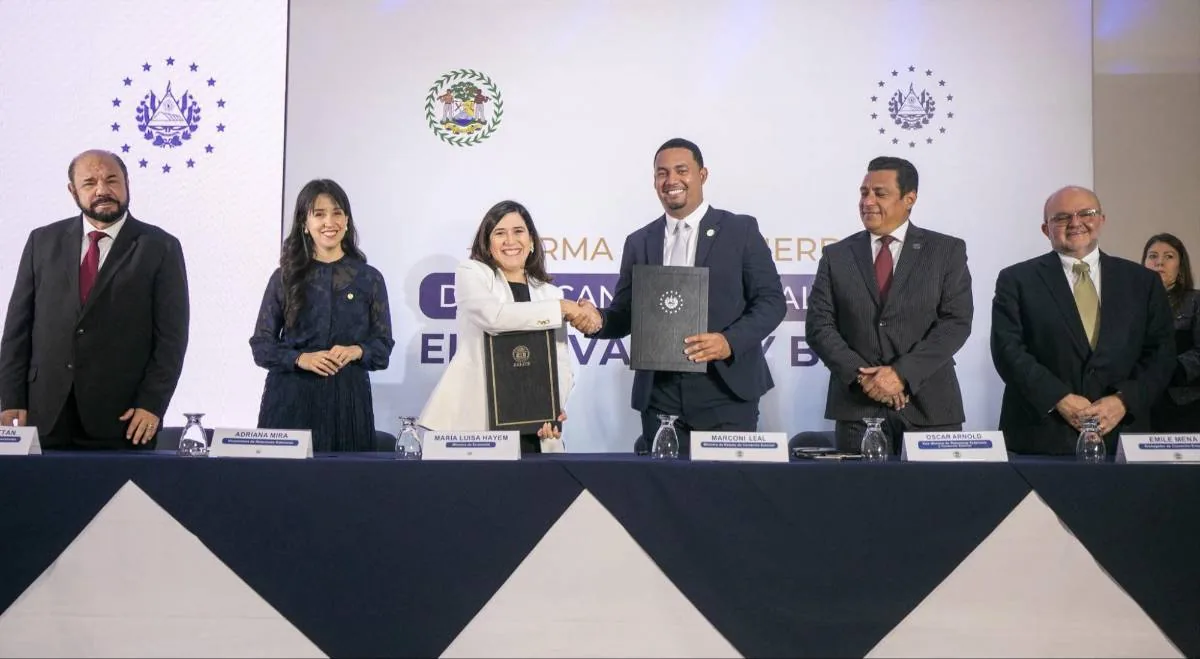 Grupo de seis ejecutivos en una ceremonia de firma de acuerdo. Dos personas centrales intercambian documentos y se estrechan la mano. Fondo con emblemas.