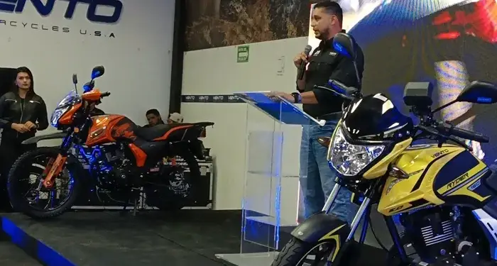 Hombre presenta nuevas motocicletas Vento en un evento de lanzamiento. Dos modelos, naranja y amarillo, en exhibición.