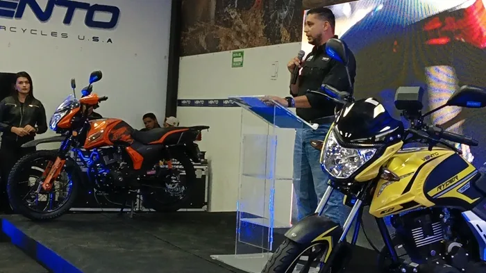 Hombre presenta nuevas motocicletas Vento en un evento de lanzamiento. Dos modelos, naranja y amarillo, en exhibición.