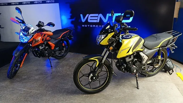 Dos motocicletas Vento, una naranja y otra amarilla, exhibidas en un evento. La naranja es tipo doble propósito, la amarilla urbana con parrilla trasera. Fondo azul con logo Vento.