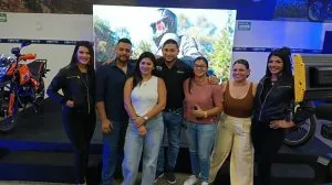 Grupo de siete profesionales, tres hombres y cuatro mujeres, posando sonrientes en un evento comercial con motocicletas y una pantalla digital de fondo.