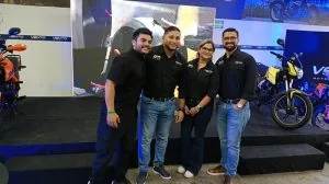 Cuatro profesionales, tres hombres y una mujer, sonríen en un evento de negocios. Visten uniformes negros y posan frente a motocicletas y pantallas con gráficos, sugiriendo una exposición comercial.