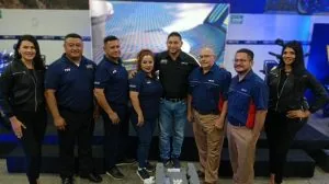 Ocho profesionales, cuatro hombres y cuatro mujeres, posando en un evento de negocios con pantalla digital de fondo. Visten ropa corporativa, incluyendo polos azules y chaquetas negras, mostrando unidad de equipo.