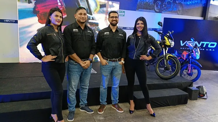 Cuatro personas, dos hombres y dos mujeres, posan frente a motocicletas Vento en una exhibición. Llevan ropa casual de negocios con logos 'AM'. El fondo muestra el logo 'VENTO MOTOCYCLES U.S.A.' y otras motos.
