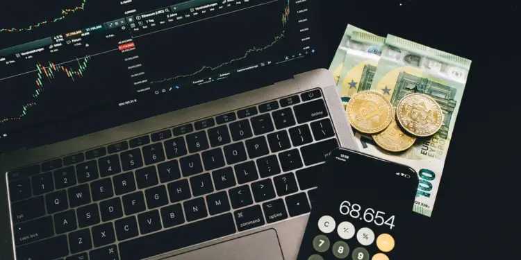 Computadora portátil con gráficos bursátiles, billetes de euro y monedas físicas de criptomonedas sobre una mesa.
