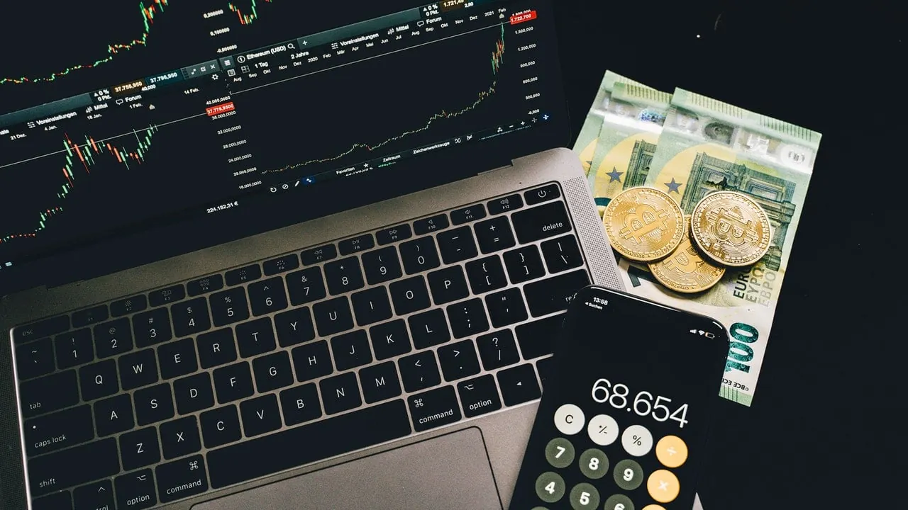 Computadora portátil con gráficos bursátiles, billetes de euro y monedas físicas de criptomonedas sobre una mesa.
