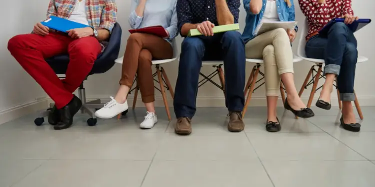 Cinco personas sentadas en fila en una sala de espera, con carpetas en sus manos, preparadas para una entrevista o reunión profesional.