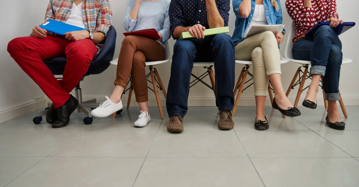 Cinco personas sentadas en fila en una sala de espera, con carpetas en sus manos, preparadas para una entrevista o reunión profesional.