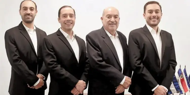 Cuatro hombres de negocios de pie vistiendo trajes oscuros y camisas blancas en una oficina.