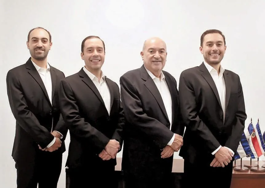 Cuatro hombres de negocios de pie vistiendo trajes oscuros y camisas blancas en una oficina.