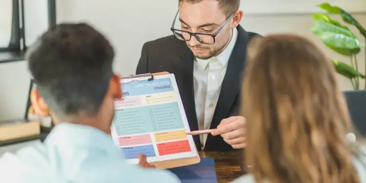 Un profesional con gafas explica un checklist de inspección de vivienda a una pareja. Se enfoca en secciones como electricidad y plomería.
