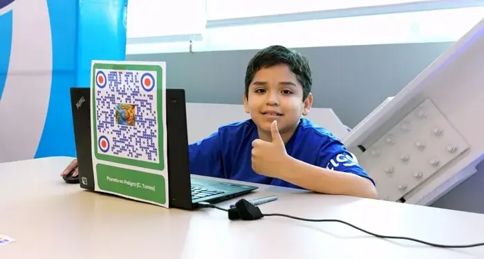 Niño sonriente con camiseta azul haciendo un pulgar arriba, sentado frente a un laptop con un código QR verde pegado en la pantalla. Un ratón y cables sobre el escritorio.