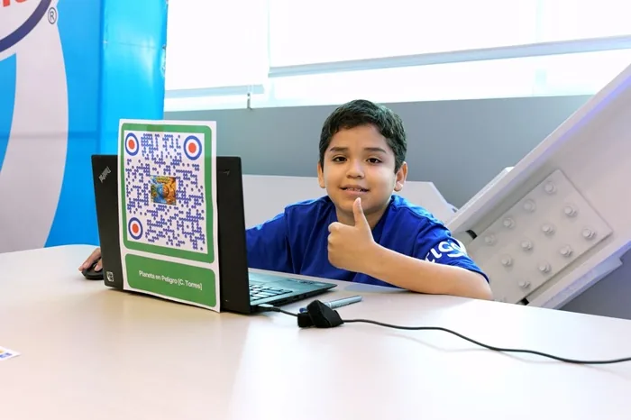 Niño sonriente con camiseta azul haciendo un pulgar arriba, sentado frente a un laptop con un código QR verde pegado en la pantalla. Un ratón y cables sobre el escritorio.