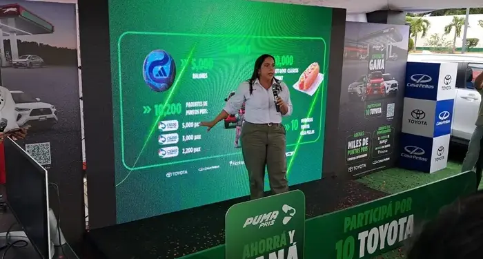 Mujer hablando con un micrófono frente a una pantalla gigante con gráficos de puntos, autos y premios.