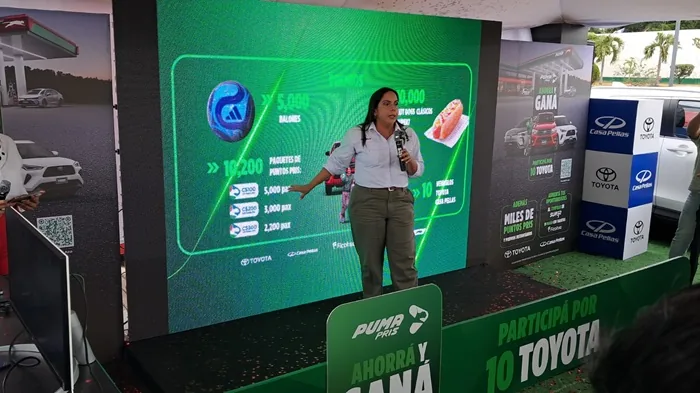 Mujer hablando con un micrófono frente a una pantalla gigante con gráficos de puntos, autos y premios.