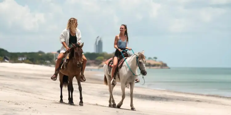 Dos mujeres jóvenes montando a caballo por la orilla de una playa arenosa junto al mar.