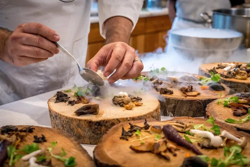 Chef profesional emplatando cuidadosamente un plato gourmet con setas y hierbas sobre rodajas de madera, rodeado de humo escénico en una cocina.