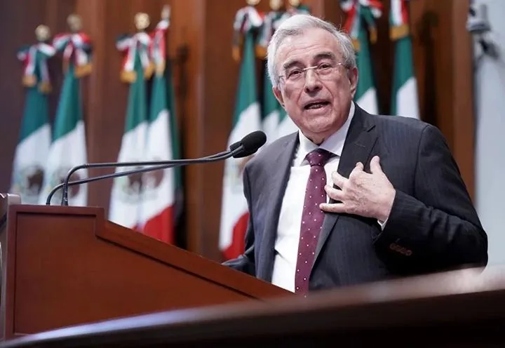 Un hombre mayor con gafas, vestido con traje y corbata, habla apasionadamente detrás de un podio de madera, con banderas nacionales al fondo.