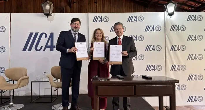 Tres profesionales sonrientes mostrando documentos firmados frente a un panel con el logotipo de IICA en un evento.