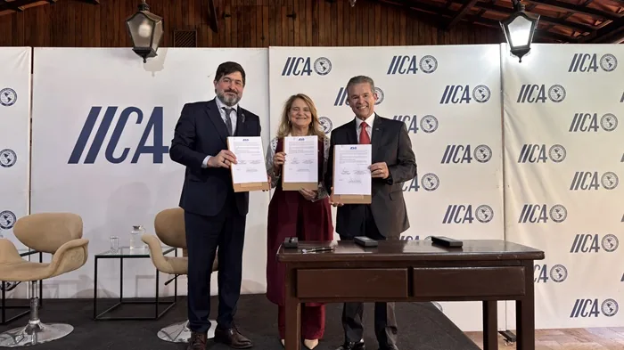 Tres profesionales sonrientes mostrando documentos firmados frente a un panel con el logotipo de IICA en un evento.
