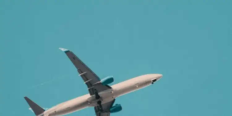 Avión comercial blanco con motores azules ascendiendo en un cielo azul claro. Vista desde abajo, simbolizando viajes y negocios.