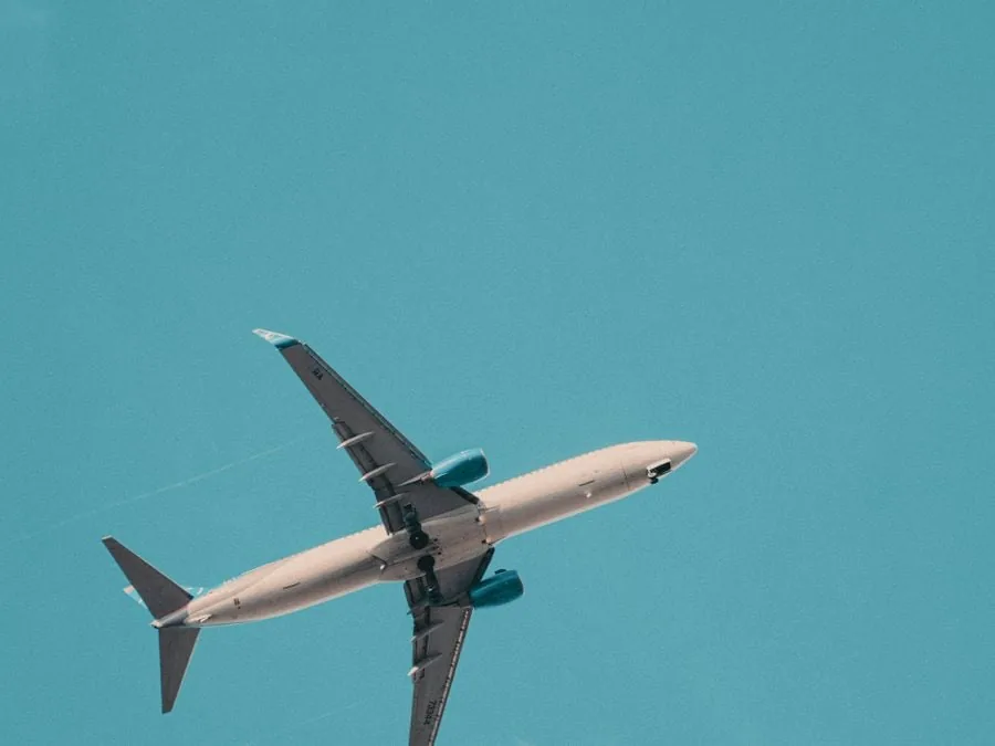 Avión comercial blanco con motores azules ascendiendo en un cielo azul claro. Vista desde abajo, simbolizando viajes y negocios.