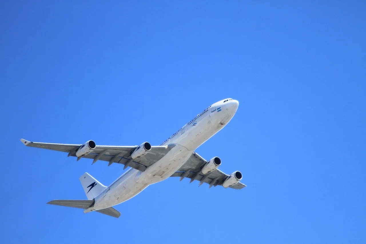 Un gran avión comercial de pasajeros con cuatro motores volando en diagonal contra un cielo azul despejado.