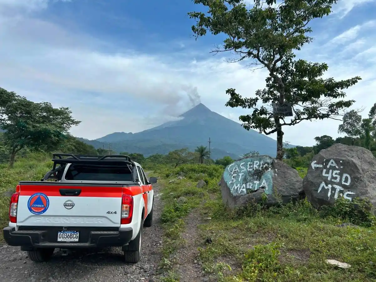 Camioneta Nissan Frontier 4x4 de CONRED Guatemala en camino rural. Volcán humeante al fondo, rocas con 'CASERIO ST MARTA' y 'A: 450 MTS'.