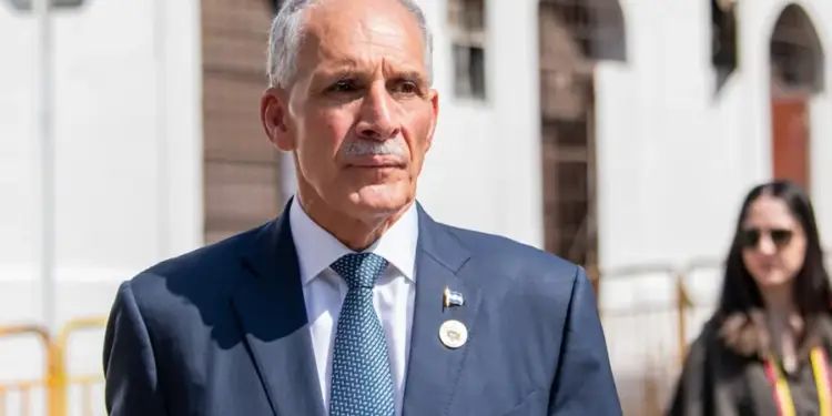 Hombre mayor de cabello canoso con traje azul, corbata y pines institucionales camina en un entorno exterior.