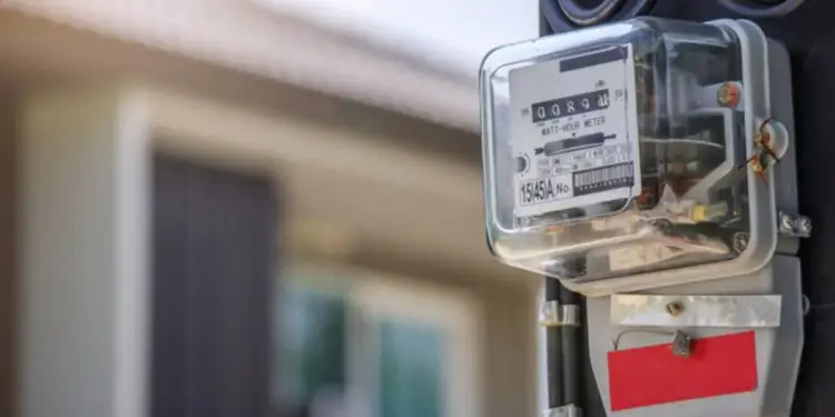 Primer plano de un contador de luz eléctrico gris con cubierta transparente, mostrando la lectura de consumo y cables negros. Al fondo, una casa borrosa.