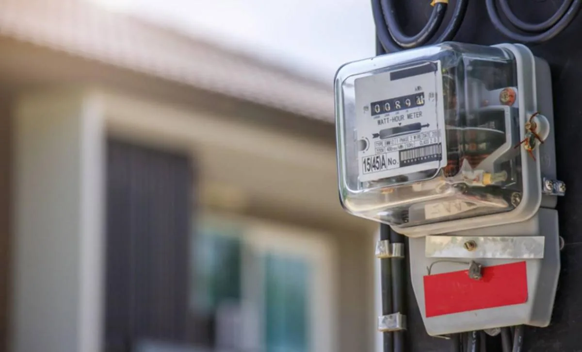 Primer plano de un contador de luz eléctrico gris con cubierta transparente, mostrando la lectura de consumo y cables negros. Al fondo, una casa borrosa.