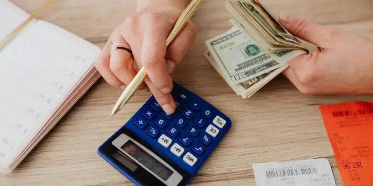 Persona utilizando una calculadora azul mientras sostiene un bolígrafo y cuenta billetes de dólares sobre una mesa.