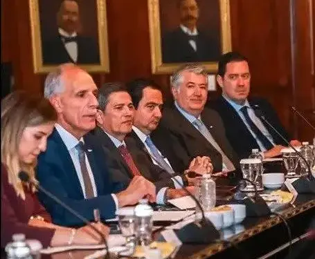 Grupo de siete profesionales, incluyendo una mujer, sentados en una mesa de reuniones con micrófonos y vasos de agua. Al fondo, dos retratos.