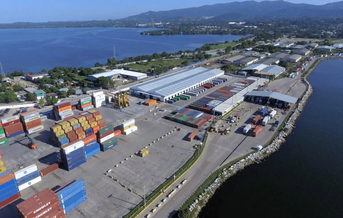 Vista aérea de una terminal portuaria con contenedores de carga apilados junto al mar y bodegas logísticas.