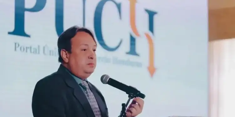 Hombre en traje oscuro y corbata hablando en un podio con micrófono. Detrás, pantalla con logo 'PUCH' y 'Portal Único de Comercio Honduras'.