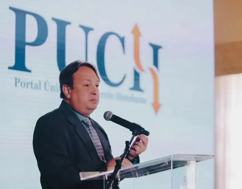 Hombre en traje oscuro y corbata hablando en un podio con micrófono. Detrás, pantalla con logo 'PUCH' y 'Portal Único de Comercio Honduras'.