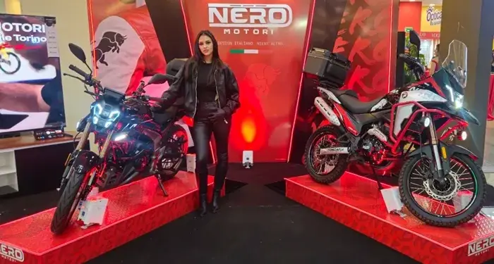Mujer en atuendo de cuero negro posa entre dos motocicletas NERO MOTORI en exhibición. Modelos de calle y aventura sobre plataformas rojas.