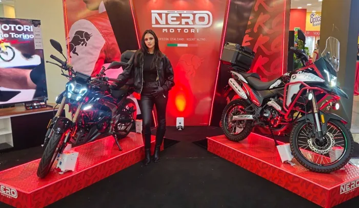 Mujer en atuendo de cuero negro posa entre dos motocicletas NERO MOTORI en exhibición. Modelos de calle y aventura sobre plataformas rojas.