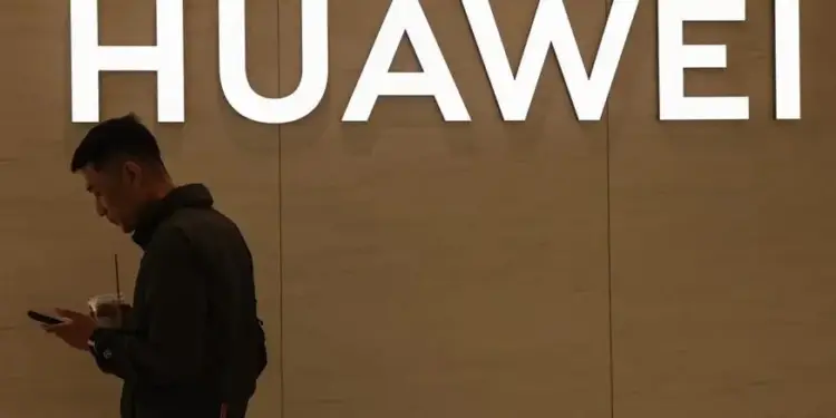 Hombre de perfil mirando su teléfono móvil frente a un cartel iluminado con el nombre de HUAWEI.