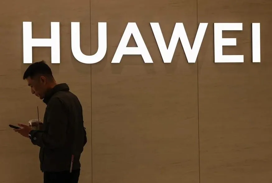 Hombre de perfil mirando su teléfono móvil frente a un cartel iluminado con el nombre de HUAWEI.