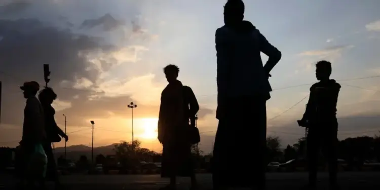 Siluetas de varias personas, principalmente hombres, de pie y caminando en una calle al atardecer. El cielo muestra tonos naranjas y azules con nubes. Se ven farolas y montañas distantes.