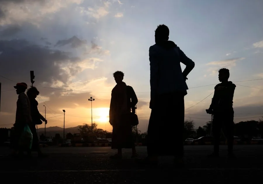 Siluetas de varias personas, principalmente hombres, de pie y caminando en una calle al atardecer. El cielo muestra tonos naranjas y azules con nubes. Se ven farolas y montañas distantes.