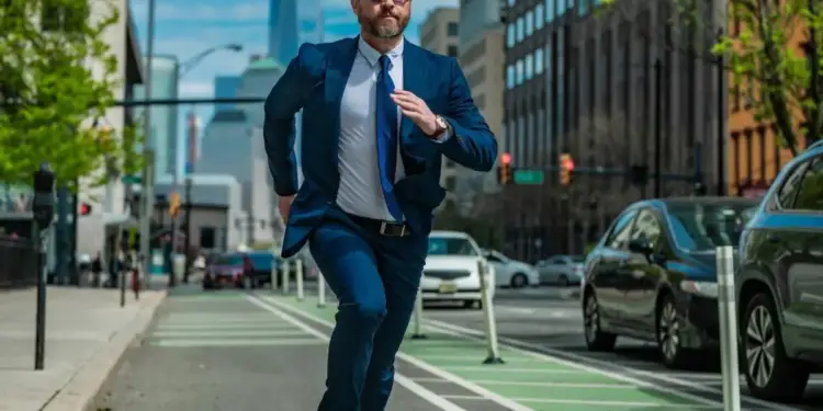 Hombre de negocios con traje azul y gafas de sol corriendo por la calle de la ciudad con rascacielos al fondo.